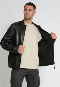 Schwarze Lederjacke mit Reißverschluss, Stehkragen und zwei seitlichen Taschen. Getragen über einem beigen Hemd und grauen Cargo-Hosen.