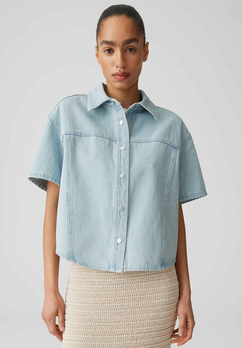 Femme aux cheveux tirés vers l'arrière portant une chemise en jean à manches courtes bleu clair boutonnée et une jupe beige en crochet sur fond gris.
