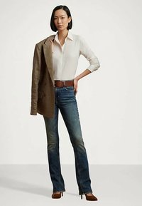 Egy nő fehér gombos inget, kék farmernadrágot, barna övet és barna sarkú cipőt visel, az egyik vállán egy barna kockás zakót tart.