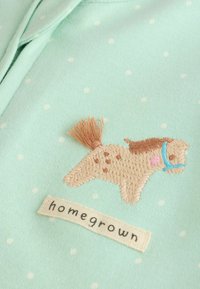 Hele sinine kangas valgete täppidega, tikitud beež hobune pruunide laigudega ja sinise jõhviga, allpool "homegrown" tekstipatch.