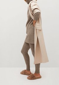 Robe-sweater beige surdimensionnée, leggings assortis et chaussons marron moelleux. Texture en tricot doux avec un manteau beige clair ceinturé drapé sur l'épaule.