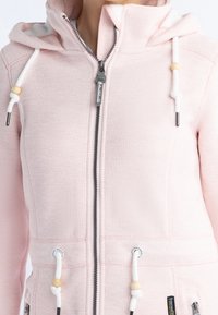 Femme portant un sweat à capuche zippé rose clair avec cordons blancs ornés de perles en bois et poches avant zippées.