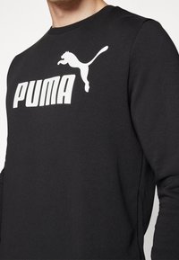 Hombre vestido con una camiseta de manga larga negra con un gran logo blanco de Puma y una gráfica de un puma saltando en el pecho.