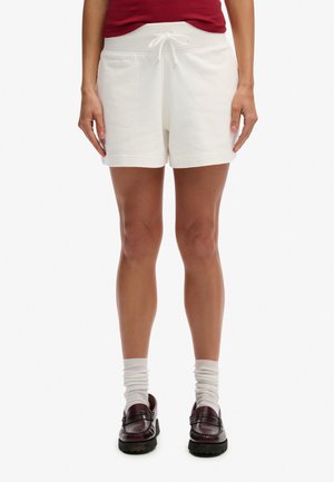 Person trägt weiße Shorts mit Kordelzug, bordeauxrotes ärmelloses Oberteil, weiße gerüschte Socken und dunkelbraune Loafers, steht vor einem weißen Hintergrund.
