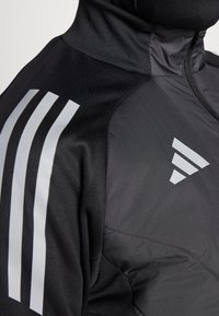 Veste de sport noire avec un col haut, un marquage argenté à trois bandes sur l'épaule et un logo triangulaire distinctif.