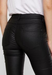 Pantalon taille haute en similicuir noir avec une texture lisse, doté de deux poches arrière et de coutures apparentes.