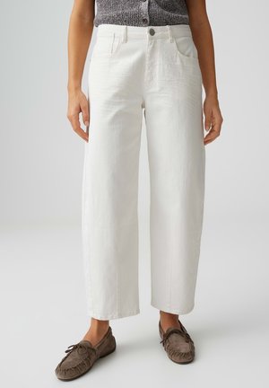 Donna indossa pantaloni bianchi a vita alta e gamba larga con tasche frontali e bottone, abbinati a un top grigio senza maniche e mocassini marroni.