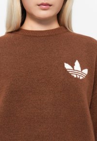 Brun stickad tröja med en vit broderad logotyp och tre bladdesigns på vänster bröstområde. Mjuk textur, ribbad halsringning.