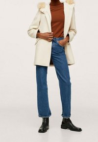 Abrigo de marfil con capucha de pelo sintético, suéter marrón de cuello alto, jeans azules y botines negros. Presenta un diseño elegante y un ajuste relajado.