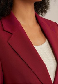 Burgundowy żakiet z klapami, wykonany z gładkiego materiału. Noszony na jasnobeżowej bluzce z prążkami, podkreślając kontrast między kolorami.