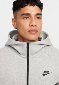 Šviesiai pilkas zip-up džemperis su juodu užtrauktuku, Nike logotipas krūtinėje ir gobtuvu. Pagamintas iš minkštos, tekstūruotos medžiagos su prigludusia forma.
