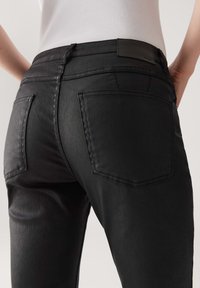 Ingrandimento di una persona che indossa jeans neri attillati e una maglietta bianca a coste, evidenziando le tasche posteriori e le cuciture.