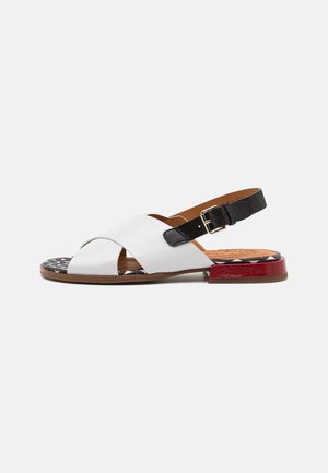 Sandale à petit talon avec des brides croisées en cuir blanc, une bride noire réglable avec boucle, une semelle à motif et un petit détail rouge au talon.