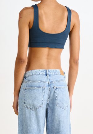 Schlanke Person in einem dunkelblauen, verkürzten Tanktop und hellblauen, hoch taillierten, weit geschnittenen Jeans, die vor einem schlichten weißen Hintergrund steht.