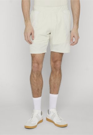Mann trägt off-white Sportshorts, weiße Sportsocken und weiße Sneaker mit Gummisohlen vor einem schlichten Hintergrund.