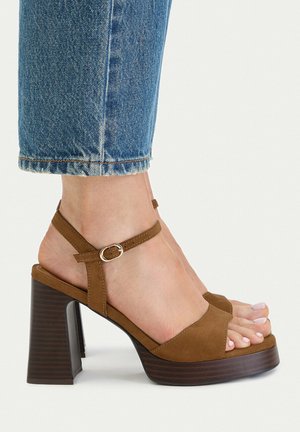 Foot wearing brown suede open-toe high-heel sandal with ankle strap and buckle, paired with cropped blue denim jeans.