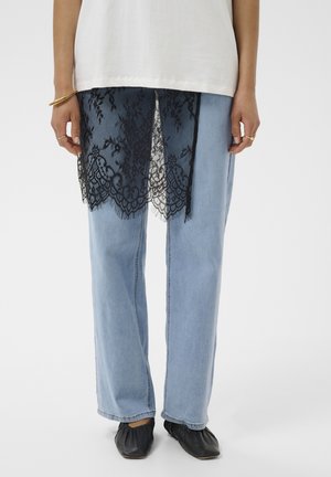 Personne portant un jean bleu clair avec un overlay en dentelle noire sur une jambe, un haut blanc, des chaussures noires, ainsi que des bagues et un bracelet dorés.