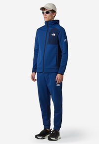 Męski niebieski strój sportowy z dopasowaną kurtką na zamek, pasującymi spodniami i czarnymi trampkami; zawiera detale z logo oraz okulary przeciwsłoneczne.