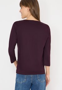 Camicia bordeaux a tre quarti con scollo rotondo. Tessuto morbido e liscio con vestibilità rilassata, abbinata a jeans in denim blu.