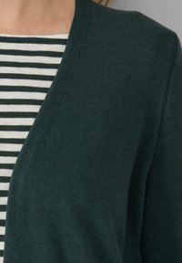 Zielony kardigan wykonany z miękkiej dzianiny, z głębokim dekoltem w serek i subtelnymi prążkowymi detalami, nałożony na bluzkę w czarno-białe paski.
