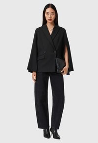 Blazer noir à double boutonnage avec des manches cape, design minimaliste ; associé à un jean noir taille haute et une pochette noire élégante.