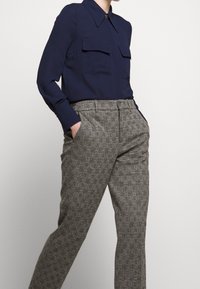 Námořnická blůza s předními kapsami, spárovaná s šedými kostkovanými kalhotami s vysokým pasem, ruce v kapsách, na jednoduchém pozadí.