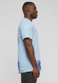 Světle modré tričko s krátkým rukávem v technice tie-dye, s tmavšími modrými akcenty na lemu a kulatým výstřihem.