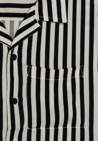 Camicia a righe bianche e nere con colletto a scollo a V, taschino frontale e bottoni neri. Il tessuto sembra morbido e leggero.