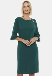 Zielona sukienka o prostym kroju, z rękawami 3/4 zakończonymi falbankami oraz dwoma ozdobnymi broszkami przy dekolcie. Gładka tekstura materiału.