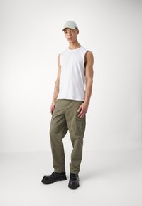 Camicetta bianca senza maniche, pantaloni cargo verde oliva e scarpe chunky nere. Il cappello è verde chiaro. Design semplice e vestibilità rilassata in tutto.