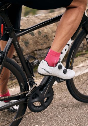 Weißer Fahrradschuhe mit schwarzen Akzenten und zwei drehbaren Dials, kombiniert mit einer rosa Socke, auf einem schwarzen Fahrrad mit sichtbarem Kettenantrieb und Pedalen.