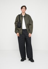 Veste bomber vert olive, t-shirt blanc, pantalons matelassés noirs et sneakers noirs. Sac bandoulière noir avec détail de logo. Fond uni.