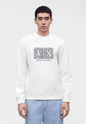 Νέος άνδρας που φοράει λευκό φούτερ Armani Exchange με μπλε στιγματισμένο λογότυπο και ανοιχτό μπλε τζιν, στέκεται μπροστά σε ένα απλό φωτεινό φόντο.