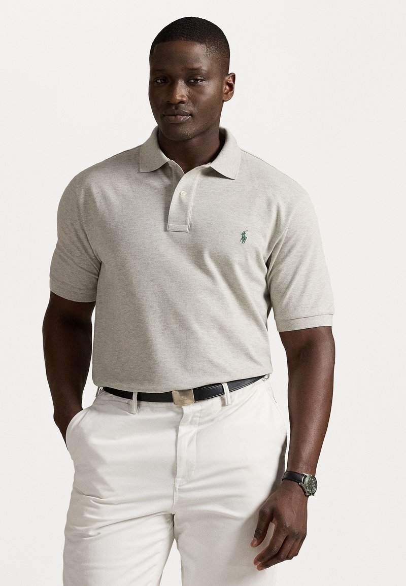 Polo shirt cinza claro com mangas curtas, com palas de dois botões, logótipo verde bordado no peito, combinado com calças brancas e cinto preto.