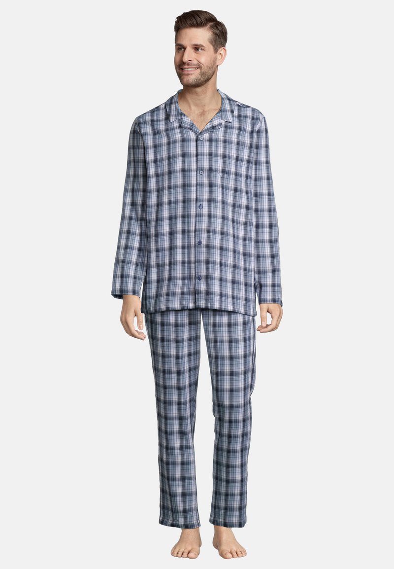 Mies seisoo paljain jaloin pukeutuneena pitkähihainen, nappikiinnitteinen yläosa ja siihen sopivat ruudulliset housut, hymyilee ja katsoo oikealle.