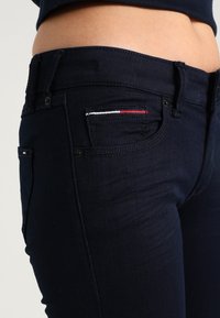 Mörkblå denimjeans med slim fit, som har en liten ficka accentuerad av röd, vit och blå sömnad. Slät textur.