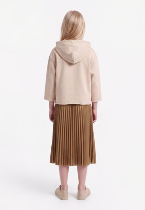 Fille aux longs cheveux blonds portant un sweat à capuche beige, une jupe plissée marron et des baskets beiges, debout de dos sur fond blanc.