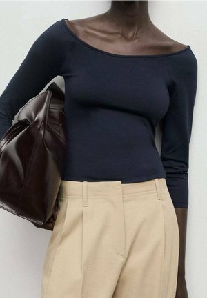 Kvinde iført en marineblå off-shoulder bluse med lange ærmer og beige højttaljede bukser, der holder en stor mørkebrun lædertaske under armen.