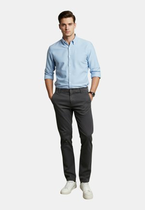 Uomo in piedi con le mani nelle tasche che indossa una camicia azzurro chiaro abbottonata, pantaloni grigio scuro, scarpe da ginnastica bianche e un orologio da polso.