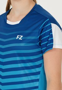 Närbild av en person som bär en blå sporttröja med ljusblå vinklar, vita ärmpaneler och en vit "FZ"-logo.