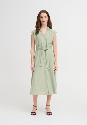 Robe chemise en lin vert avec des manches courtes, une fermeture à boutons sur le devant et une ceinture assortie à la taille ; le modèle porte des sandales noires à lanières.