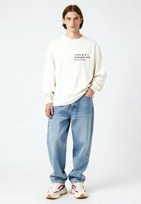 Jeune homme aux cheveux bruns portant une chemise blanche à manches longues avec un texte, un jean large bleu clair et des baskets blanches avec des accents rouges et bleus.