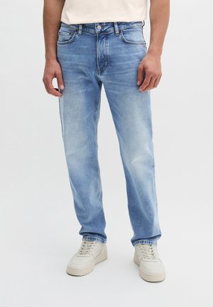Mann trägt hellblaue ausgewaschene Jeans und weiße Sneakers und steht vor einem schlichten Hintergrund.