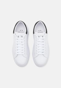 Sneaker bianche in pelle con un accento nero sul tallone, dotate di lacci piatti, dettagli cuciti e loghi sulla linguetta e sul lato.
