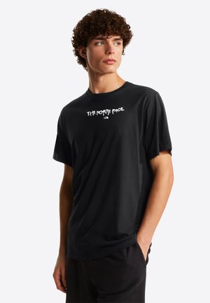 Ung mand med krøllet hår iført en sort T-shirt med "The North Face"-logo, stående med den ene hånd i lommen og kigger til venstre.