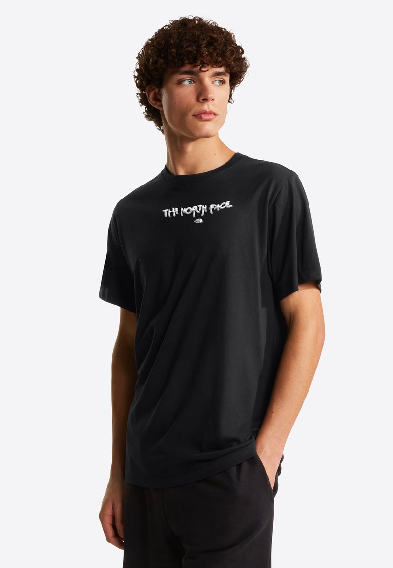 Young man with curly hair wearing a black T-shirt with "The North Face" logo, standing with one hand in pocket, looking to his left.