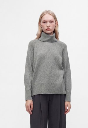 Jeune personne aux longs cheveux blonds portant un pull à col roulé gris et un pantalon large foncé, debout devant un fond blanc uni.