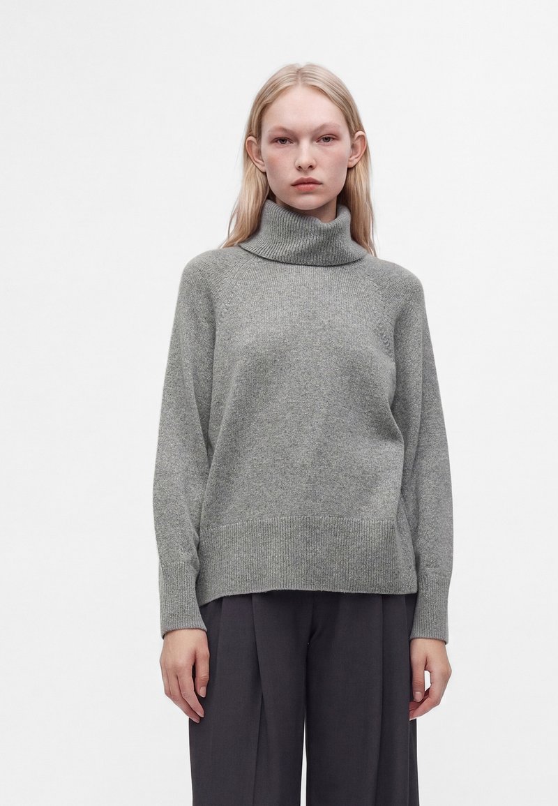 Jeune personne aux longs cheveux blonds portant un pull à col roulé gris et un pantalon large foncé, debout devant un fond blanc uni.