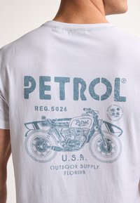 Bílá bavlněná tričko s modrou vintage grafikou motocyklu a slovem "PETROL" v opotřebovaném písmu, s dalším textem níže.