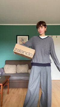 Brauner Karton mit "zalando" in großen grauen Buchstaben bedruckt. Gestreifter grau-weißer Pullover, weite graue Hose, Innenraum.